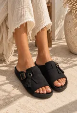 Sandalias planas color beige, fabricadas en gamuza, con tira ancha en el empeine, hebilla metálica de ajuste y detalle de flecos colgantes con argolla.