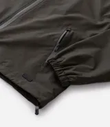 Campera rompevientos color tostado, con capucha ajustable mediante cordones, cierre frontal completo y puños elásticos. Presenta un diseño minimalista y funcional, ideal para protección contra el viento.