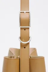 Bolso de hombro tipo cubo de estructura rígida, color marrón oscuro, confeccionado en piel vacuna. Posee asa regulable con hebilla metálica y cierre de cordón en la parte superior. Lleva el logo metálico de Bimba y Lola en la parte frontal inferior.