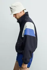 Chaqueta técnica de corte relajado con cuello subido y cierre frontal de cremallera bidireccional. Presenta un diseño color block en tonos azul marino, blanco, rojo y azul eléctrico. Incluye bolsillos frontales de vivo, puños y ruedo elásticos, y forro interior de red.