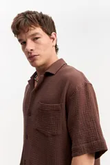 Camisa de manga corta de muselina color marrón, con cuello abierto, cierre frontal de botones y un bolsillo ojal en el pecho.