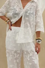Pantalón largo blanco de corte recto y holgado, confeccionado en tejido semitransparente con diseño bordado de motivos vegetales. Cuenta con cintura elástica ajustable con cordón.