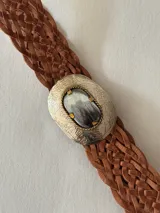 Cinto trenzado de cuero color marrón, con hebilla metálica ovalada grabada y detalle central de piedra natural.