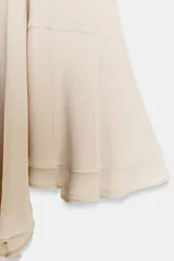 Top tipo capa de color beige, con cuello subido y cierre posterior con botones ocultos. Presenta un diseño fluido con bajo asimétrico y mangas acampanadas.