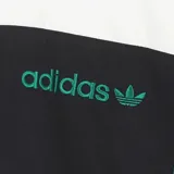 Campera deportiva Adidas con diseño color block en blanco, negro y verde. Presenta cuello alto, cierre frontal completo y las icónicas tres tiras de la marca en los laterales en color verde. El logo de Adidas está bordado en el pecho.