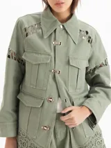 Chaqueta de gabardina liviana color verde militar, con silueta recta, largo por debajo de la cadera y cuello de corderoy. Presenta detalles de encaje en hombros, mangas y ruedo, múltiples bolsillos con solapa y cierre frontal con broches metálicos.