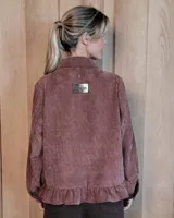 Sobrecamisa de pana color marrón oscuro, con cuello camisero, cierre frontal de botones y detalle de volado en el ruedo. Presenta mangas largas con puños y bolsillos frontales aplicados.