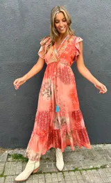 Vestido largo anaranjado con estampado estilo paisley y geométrico, escote en V con bordados multicolor, mangas cortas con volados, cordón ajustable en la cintura con borlas turquesas y falda escalonada.