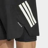 Short deportivo Adidas 2 en 1 con calza interior integrada, diseñado para running. Presenta un diseño en color negro con las icónicas tres tiras de la marca en tono claro sobre el muslo y logo en la cintura. Incluye cintura elástica con cordón de ajuste y bolsillos laterales con cierre.
