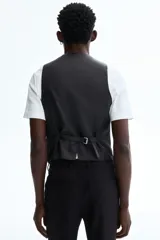 Chaleco de traje formal con cierre frontal de cinco botones y dos bolsillos delanteros ribeteados. Presenta un corte ajustado, forro interior y espalda con tejido brillante y trabilla ajustable.