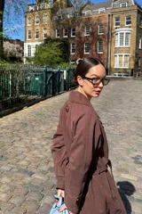 Trench coat de color marrón, con cuello solapa, cierre frontal y cinturón ajustable a la cintura. Presenta un diseño clásico con costuras estructuradas y una etiqueta pequeña en la parte superior de la espalda.
