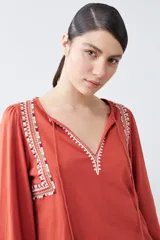 Remera blanca de manga larga con detalles bordados en tonos azules y verdes en el pecho y hombros. Presenta un escote en V con cordones ajustables terminados en borlas y puños elásticos.