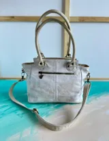 Cartera de cuero blanca y dorada con solapa texturizada tipo cocodrilo y detalles pintados a mano. Cuenta con doble asa de mano, correa ajustable, herrajes metálicos y cierre superior.