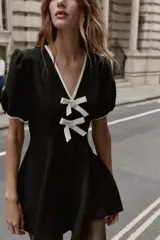 Vestido corto negro con escote en V ribeteado en blanco. Presenta mangas cortas abullonadas y dos lazos blancos decorativos en el centro del pecho.