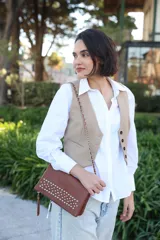 Cartera tipo sobre color tabaco, con diseño rectangular y frente decorado con tachas metálicas plateadas. Incluye una correa de cadena metálica entrelazada con cuero y un detalle de borla lateral.