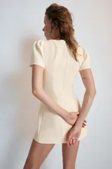 Vestido mono corto de color beige claro, con cuello redondo y mangas cortas abullonadas. Cuenta con forro interior tipo shorts y cierre en la espalda con cremallera oculta.