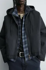 Cazadora tipo bomber de denim color negro con efecto lavado, corte boxy fit, cierre frontal de cremallera, capucha ajustable con cordón y bolsillos laterales de vivo.