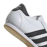 Championes Adidas modelo Taekwondo Lace, de diseño minimalista en color blanco con las icónicas tres tiras laterales en negro y detalle en el talón. Presenta una puntera reforzada con costuras decorativas y suela de goma delgada.