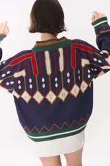 Campera tipo bomber tejida con diseño jacquard geométrico en tonos azul marino, blanco, rojo y verde. Presenta cierre frontal completo, cuello, puños y ruedo acanalados en color verde y blanco, con corte oversized y hombros caídos.