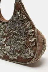 Bolso de mano mini con cuerpo de efecto aterciopelado en color marrón, adornado con abalorios y lentejuelas que forman un estampado floral. Cuenta con doble asa de mano rígida decorada con cuentas y cierre magnético.