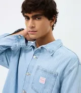 Camisa masculina de corte boxy confeccionada en denim de algodón, con cuello camisero, mangas largas, cierre frontal con botones y bolsillo funcional en el pecho.