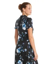 Vestido midi de seda fría con estampado floral en tonos azules sobre fondo negro. Presenta escote en V con cierre frontal de botones, mangas cortas con terminación fruncida, corte en la cintura con lazo de ajuste y falda plisada.