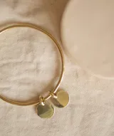 Pulsera rígida dorada tipo esclava, elaborada en bronce, con dos dijes circulares colgantes.