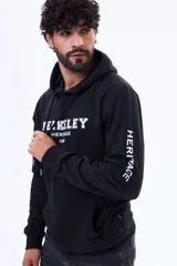 Buzo tipo canguro de felpa color negro, con capucha ajustable mediante cordones, bolsillo frontal tipo canguro y estampa frontal con texto universitario.