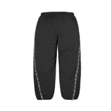 Pantalón deportivo Supreme de color negro, confeccionado en poliéster. Presenta una banda lateral con el logo de la marca repetido y el logo de Supreme bordado en la parte superior delantera. Cuenta con cintura elástica y puños ajustados en los tobillos.