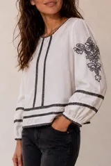 Blusa blanca de manga larga con detalles de bordados florales en tono grisáceo y bandas decorativas en el frente y mangas. Cuenta con cuello redondo y puños elásticos.