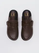 Mocasines tipo mule de cuero marrón con textura granulada, detalle de tira con hebilla metálica y ojales decorativos en el empeine.