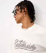 Remera boxy de algodón color blanco crudo, con cuello redondo, mangas cortas y estampa frontal en tipografía gótica con efecto de alambre de púa.