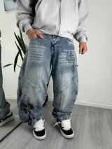 Pantalón jeans estilo baggy, color azul con lavado acid wash y efecto desgastado. Presenta diseño amplio, costuras diagonales en las piernas y estampados gráficos en estilo gótico/cyberpunk en la parte frontal. Cierre frontal con botones metálicos a la vista.