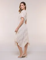 Vestido midi blanco de estilo romántico confeccionado en poliéster con elastano, con detalles de encaje floral. Cuenta con escote cruzado en V, mangas cortas acampanadas, cintura elastizada y falda evasé con terminación asimétrica en picos.