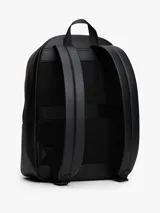 Mochila urbana de diseño minimalista confeccionada en poliuretano negro. Presenta un compartimento principal amplio con cierre, un bolsillo frontal con textura acanalada y el logo de la bandera de la marca en la esquina inferior. Incluye correas de hombro acolchadas y ajustables, además de un asa superior de mano.