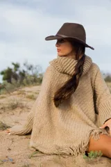 Poncho de tejido de punto grueso en color beige, con cuello alto tipo polera, mangas anchas y terminación con flecos en el ruedo.