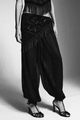 Pantalón bombacho negro semitransparente de tiro medio y cintura elástica. Tiene un pareo superpuesto ajustable con lazada lateral, decorado con abalorios, lentejuelas y flecos plateados. El bajo es elástico.