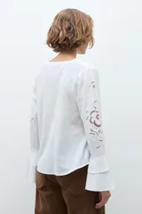 Blusa blanca de manga larga con diseño calado y bordado floral. Presenta un escote en V con detalle de volados frontales y mangas acampanadas con terminación en volado.