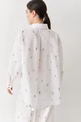 Camisa de lino blanca con estampado de figuras deportivas pequeñas y detalles de costuras en color rojo. Presenta cuello clásico, cierre frontal con botones de madera, mangas largas con puños abotonados y un bolsillo frontal aplicado.