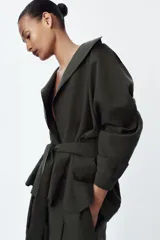 Trench corto de corte fluido confeccionado en mezcla de lyocell, color verde militar. Presenta cuello solapa, cierre frontal cruzado con botón, mangas largas con hombro caído y puños con trabilla. Incluye bolsillos delanteros de plastrón y cinturón de lazada en el mismo tejido.