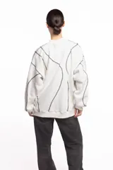 Buzo blanco de manga larga con diseño holgado, cuello redondo y costuras externas visibles en contraste. Cuenta con una etiqueta decorativa en el frente inferior.