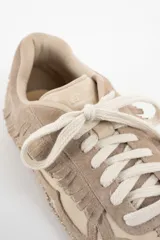 Zapatilla de cuero legítimo en tonos beige y crema, con diseño de flecos en el lateral y costuras decorativas visibles en el contorno. Presenta cordones al tono y suela plana.
