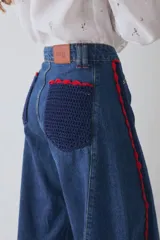 Jean de corte wide leg en color azul denim, con detalle de apliques de crochet en color rojo y azul a lo largo de las costuras laterales frontales. Presenta ruedo deshilachado y cierre frontal con botón metálico.