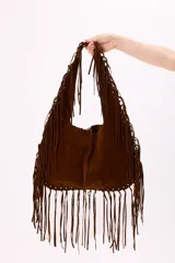 Bolso estilo hobo confeccionado en símil gamuza color marrón, caracterizado por su diseño amplio y decorado con flecos largos en todo el contorno y las asas.