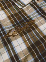 Camisa de manga corta con corte híbrido boxy fit/oversize, confeccionada en algodón. Disponible en tres diseños: a cuadros marrones, a rayas blancas y negras, y a rayas burdeos y blancas. Presenta bordados de diseño propio de la marca.