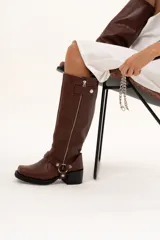 Bota de caña alta estilo biker confeccionada en cuero color chocolate. Presenta un cierre metálico lateral que recorre toda la caña, taco cuadrado y plataforma. Incluye un sistema de estribo y cadena metálica desmontable en la zona del tobillo, permitiendo diversas formas de uso.