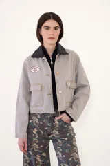 Campera corta de jean color visón, con cuello desmontable de tela engomada, cierre frontal y bolsillos plaqué con solapa en el pecho. Cuenta con un parche bordado en el delantero.