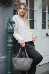 Cartera tipo tote de diseño curvo, confeccionada en cuero sintético color gris. Presenta asas regulables con hebillas metálicas y un aplique triangular metálico con el logo de la marca en el frente.