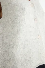 Chaleco de punto con escote en V y cierre frontal de botones. Presenta un diseño de corte recto y textura jaspeada en tono crema.