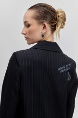 Blazer negro de rayas finas blancas, con solapas, cierre de botón frontal y broches decorativos en la solapa.